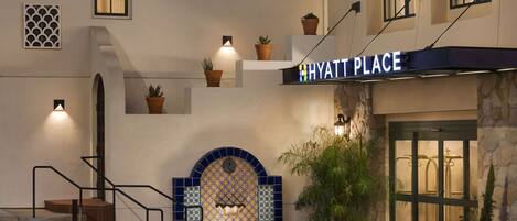 A hotel entrance with a sign reading "HYATT PLACE," a tiled wall with a geometric pattern, and potted plants.
