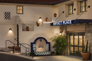 A hotel entrance with a sign reading "HYATT PLACE," a tiled wall with a geometric pattern, and potted plants.