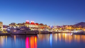 Un puerto deportivo con yates amarrados, un hotel con letreros iluminados y un paisaje costero al anochecer.