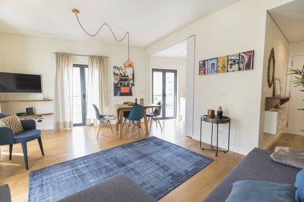 A modern living room with a sofa, a TV, a dining area with chairs, and a large window.