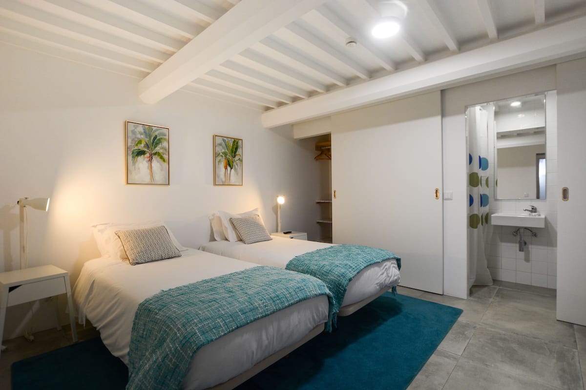 A bedroom with two beds, a nightstand, a lamp, and a bathroom with a sink and mirror.