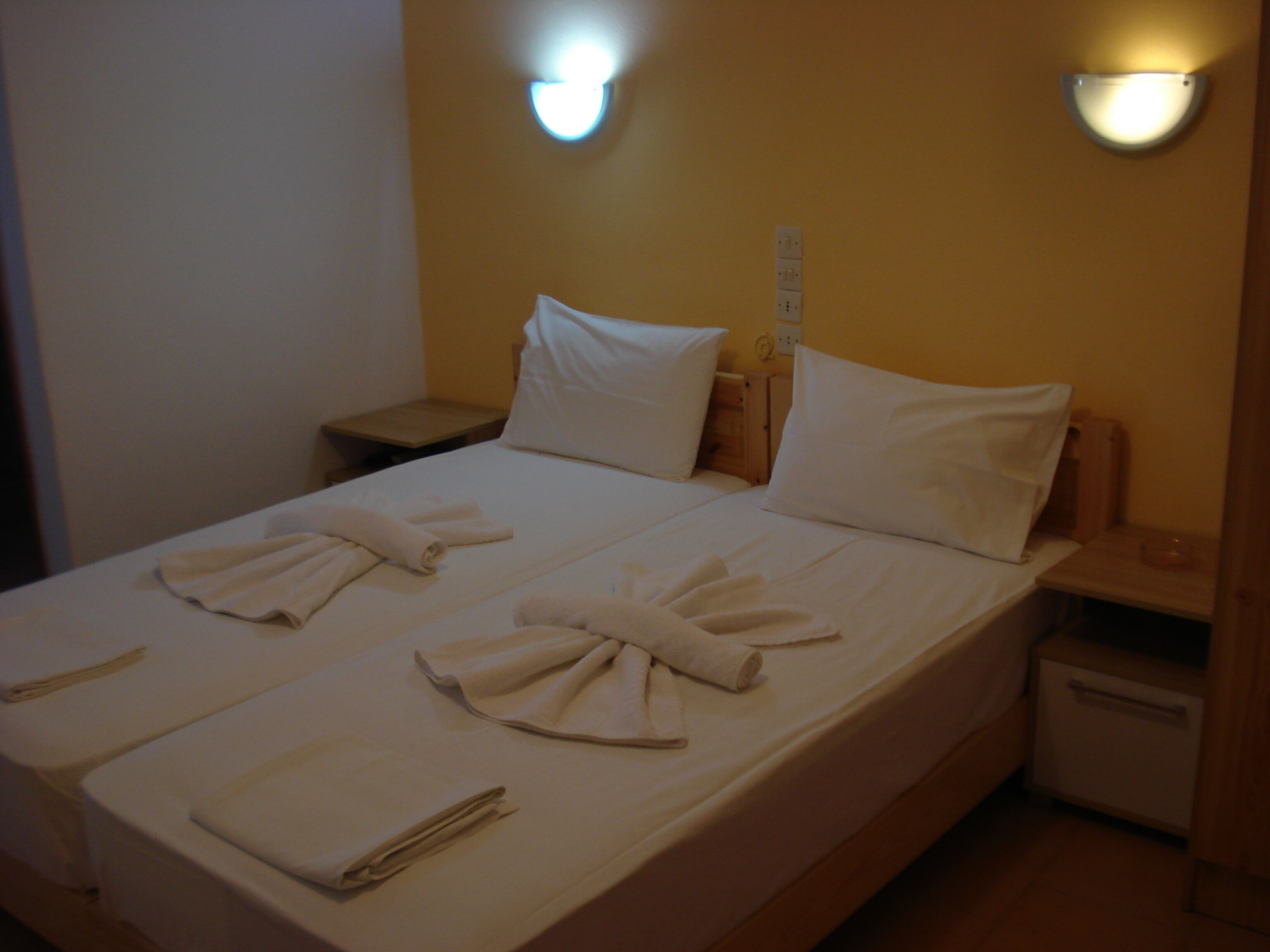 A neatly made bed with towels arranged in a bow shape, a bedside table, and a nightstand with a lamp.