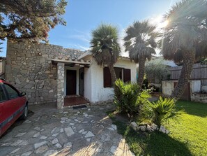 A stone house with a paved entrance, a red car parked outside, and a small garden with palm trees.
