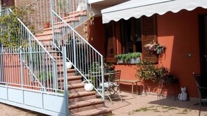 Un edificio de dos pisos con fachada de ladrillo, un balcón con plantas, una escalera y asientos al aire libre.