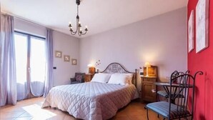 Un dormitorio con una cama grande, una araña, una mesita de noche con lámpara, una silla y una pared roja decorada con cuadros.