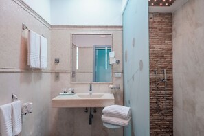 A bathroom with a beige sink, a mirror, a towel rack, and a shower area with a stone wall. - Hotel Trecento (Rome)