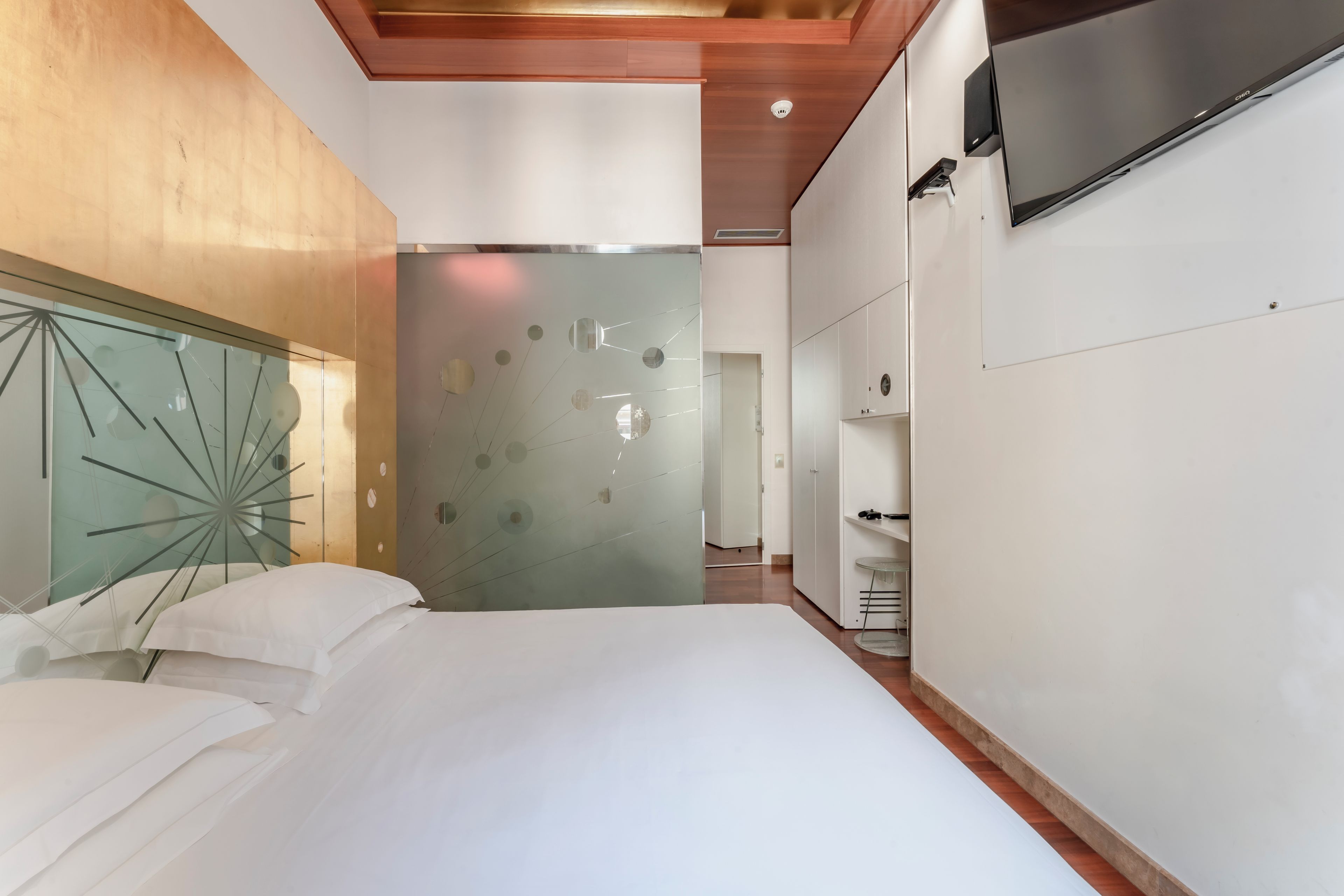 A modern hotel room with a large bed, a glass partition wall, and a mounted TV.