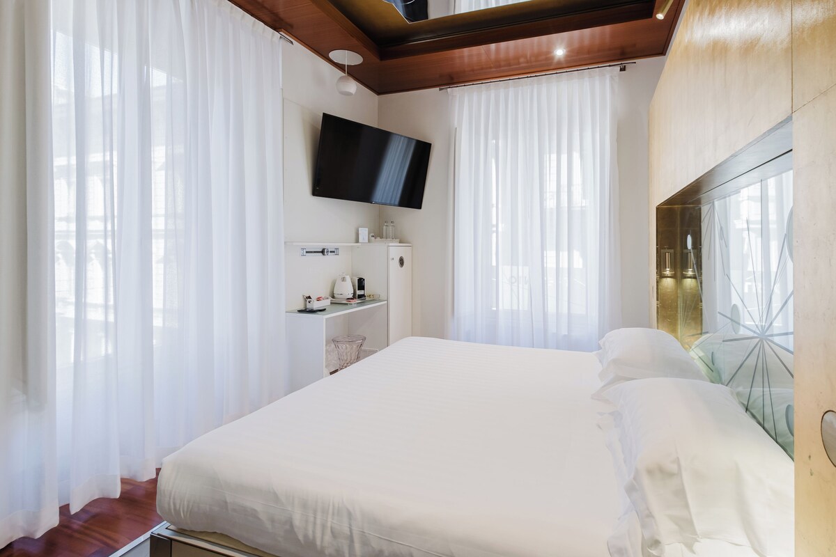 Habitació d'hotel moderna amb un llit gran, una televisió de pantalla plana penjada a la paret i una separació de vidre amb un marc metàl·lic decoratiu.