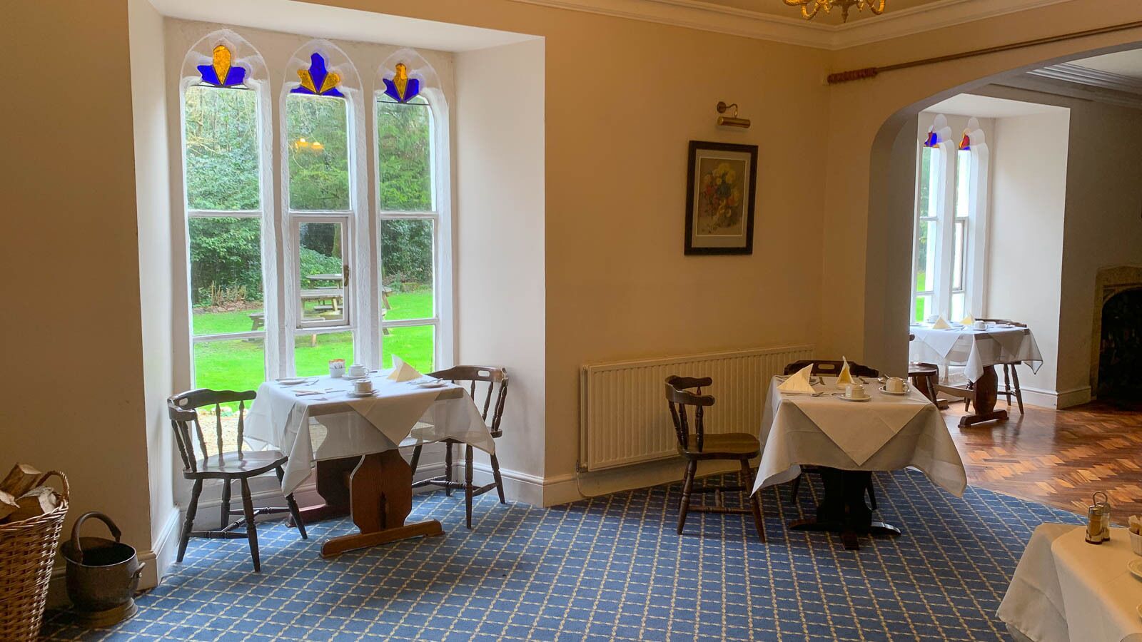 Ein Esszimmer mit zwei Tischen, Stühlen und Blick auf einen Garten durch große Fenster.