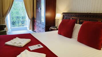 A hotel room with a large bed, red pillows, a wooden headboard, a nightstand with a lamp, and a view of greenery through the window.