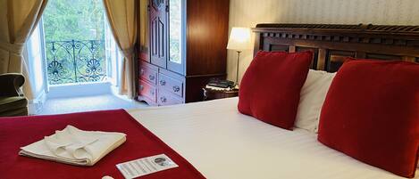 Une chambre d’hôtel avec un grand lit, des oreillers rouges, une tête de lit en bois, une table de chevet avec une lampe et une vue sur la verdure par la fenêtre.