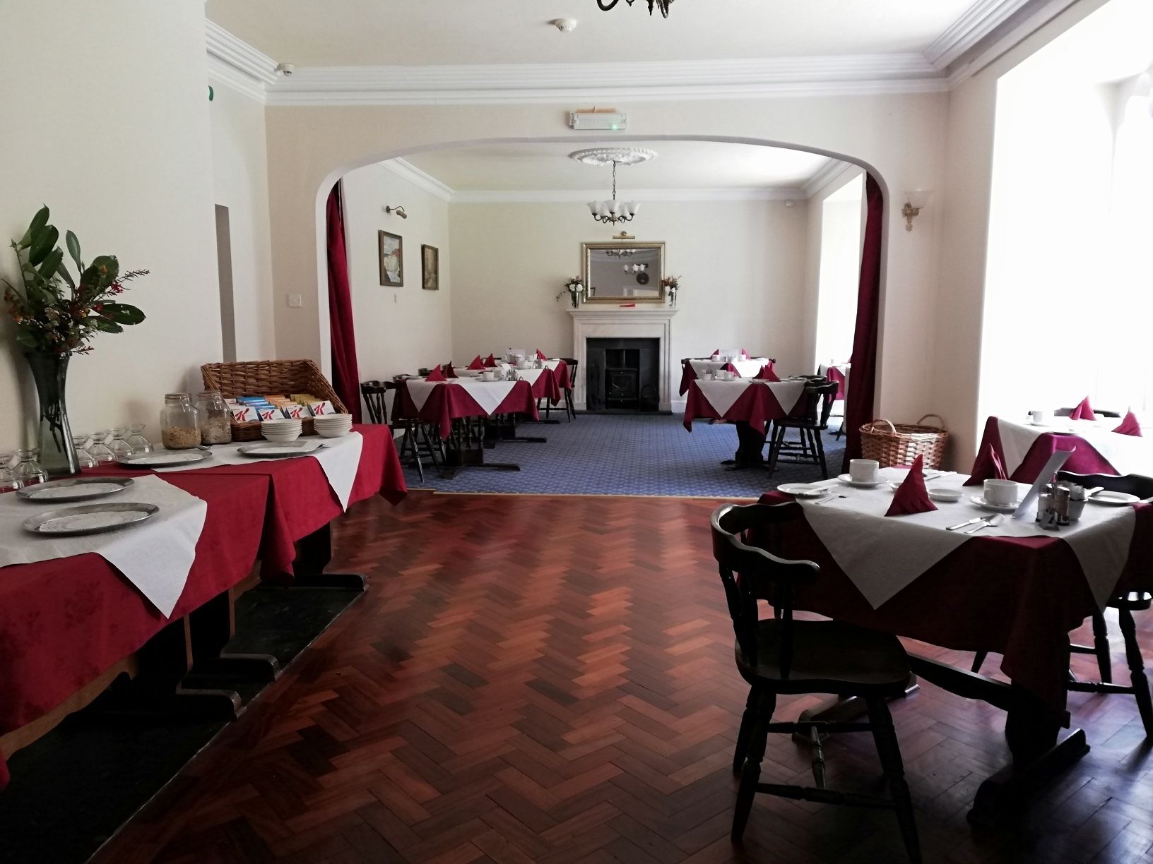 A dining hall with multiple tables set for service, a fireplace, and a large arched doorway.