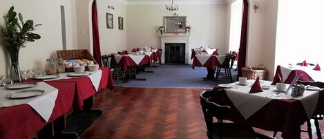 A dining hall with multiple tables set for service, a fireplace, and a large arched doorway.