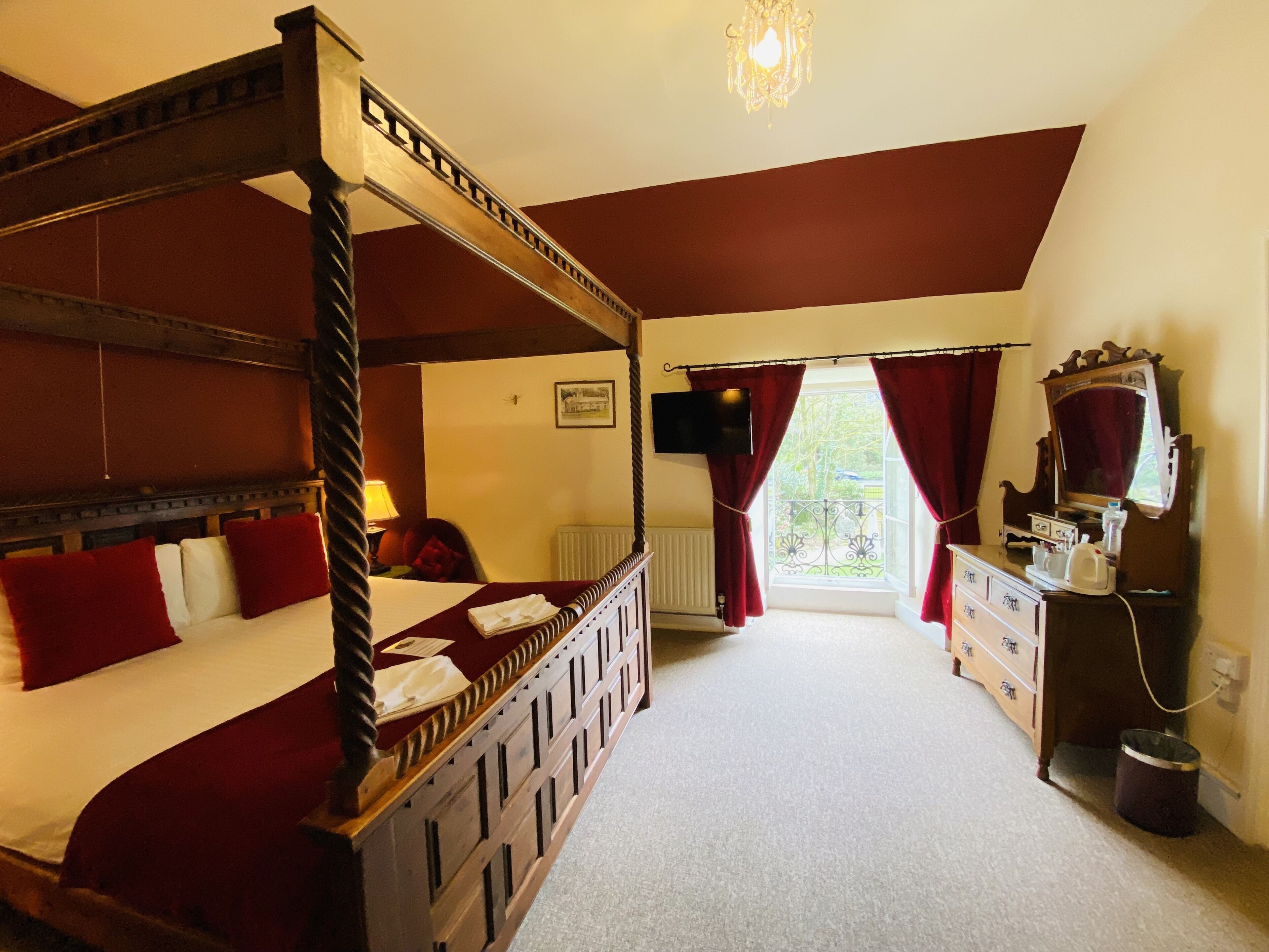 A four-poster bed with red bedding, a wooden dresser with a mirror, and a window with red curtains.