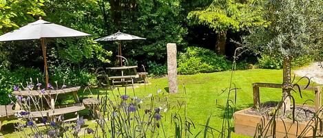 Un jardín con mesas de picnic y sombrillas, rodeado de exuberante follaje y una construcción de piedra.