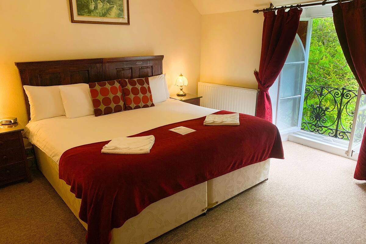A neatly made bed with a red blanket and two towels, a bedside lamp, and a window with curtains.