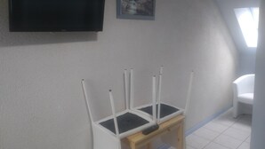 A room with a television mounted on the wall, a small table with a remote control, and a window with curtains. - Auberge du Tranchoir (Nevoy)