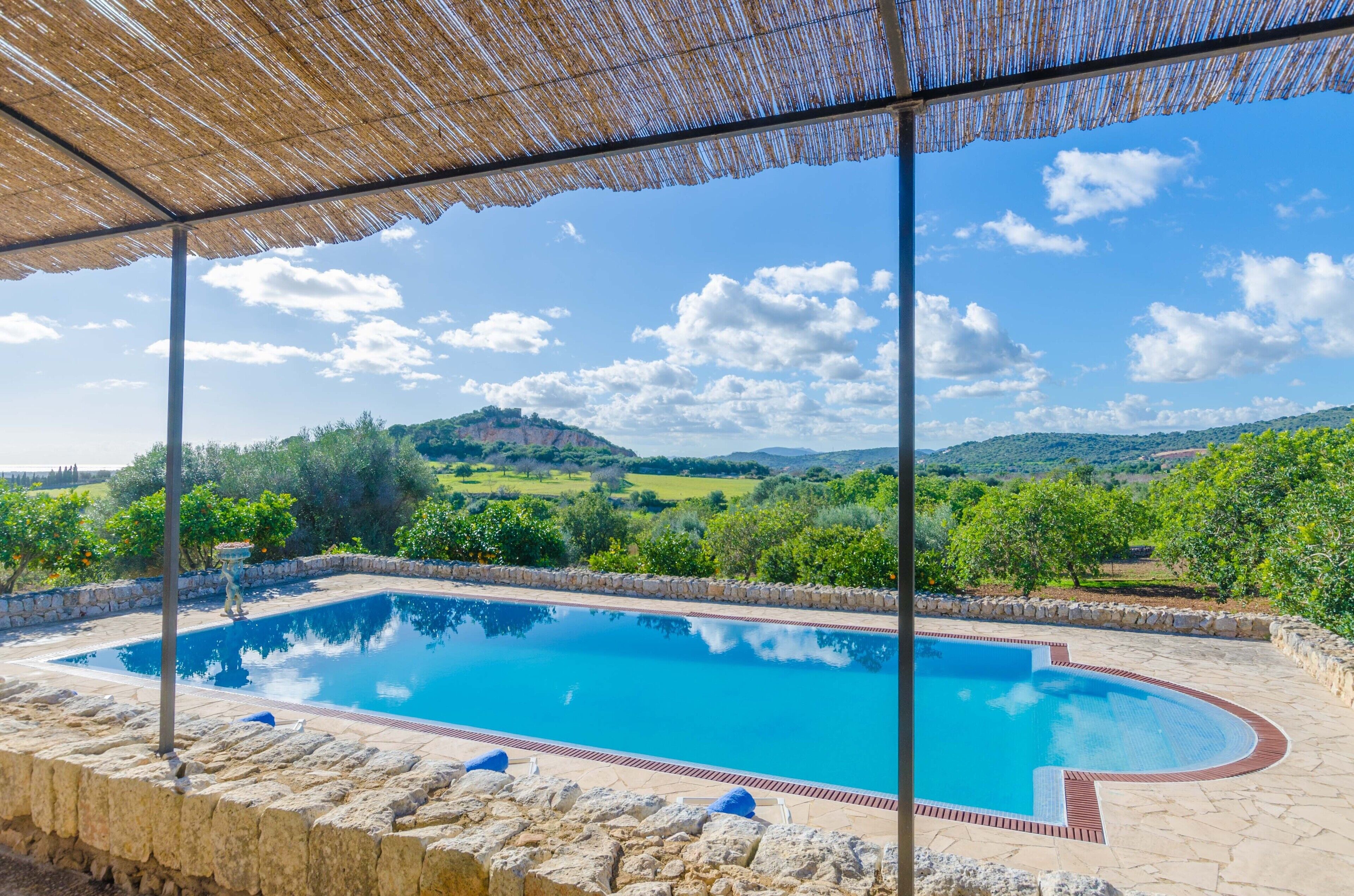 A swimming pool with a stone border, surrounded by greenery and hills in the background.