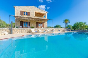 Una casa de piedra de dos pisos con piscina, sillas de descanso y un porche cubierto.