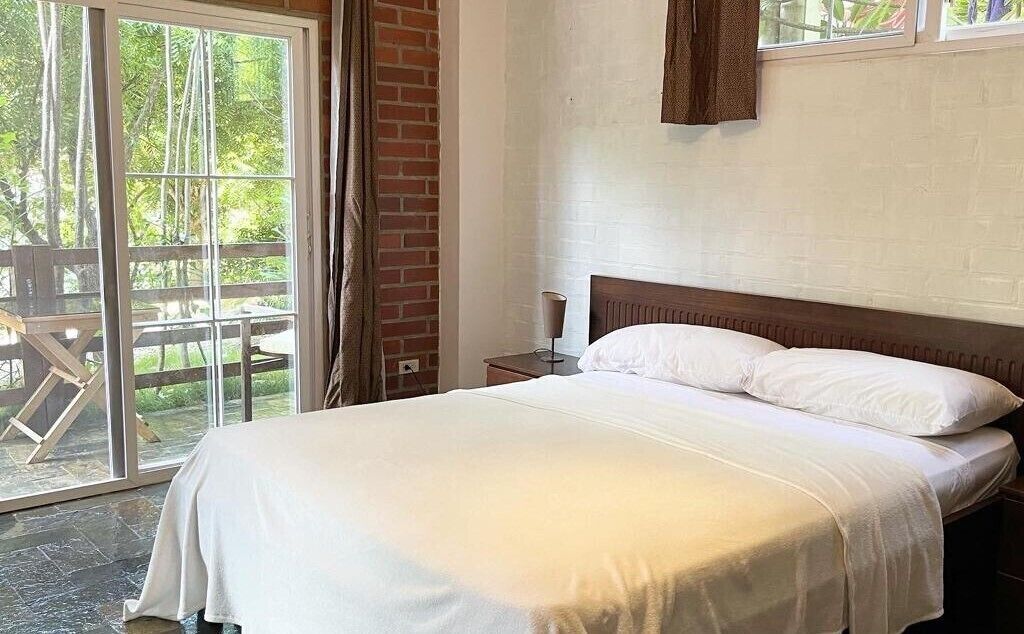 A bedroom with a large bed, a wooden headboard, a nightstand, and a window with a view of greenery.