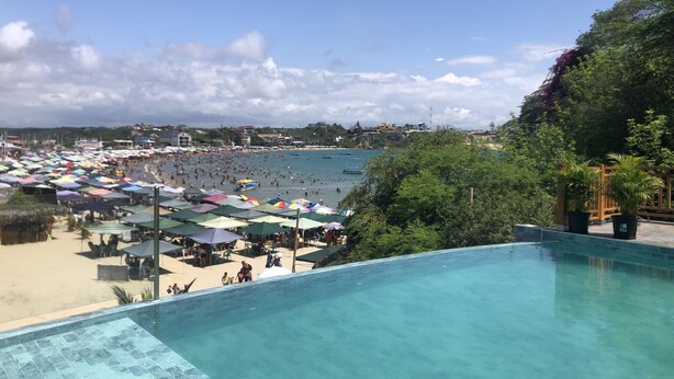Une vue depuis la piscine donnant sur une plage bondée, avec des parasols et de nombreuses personnes.