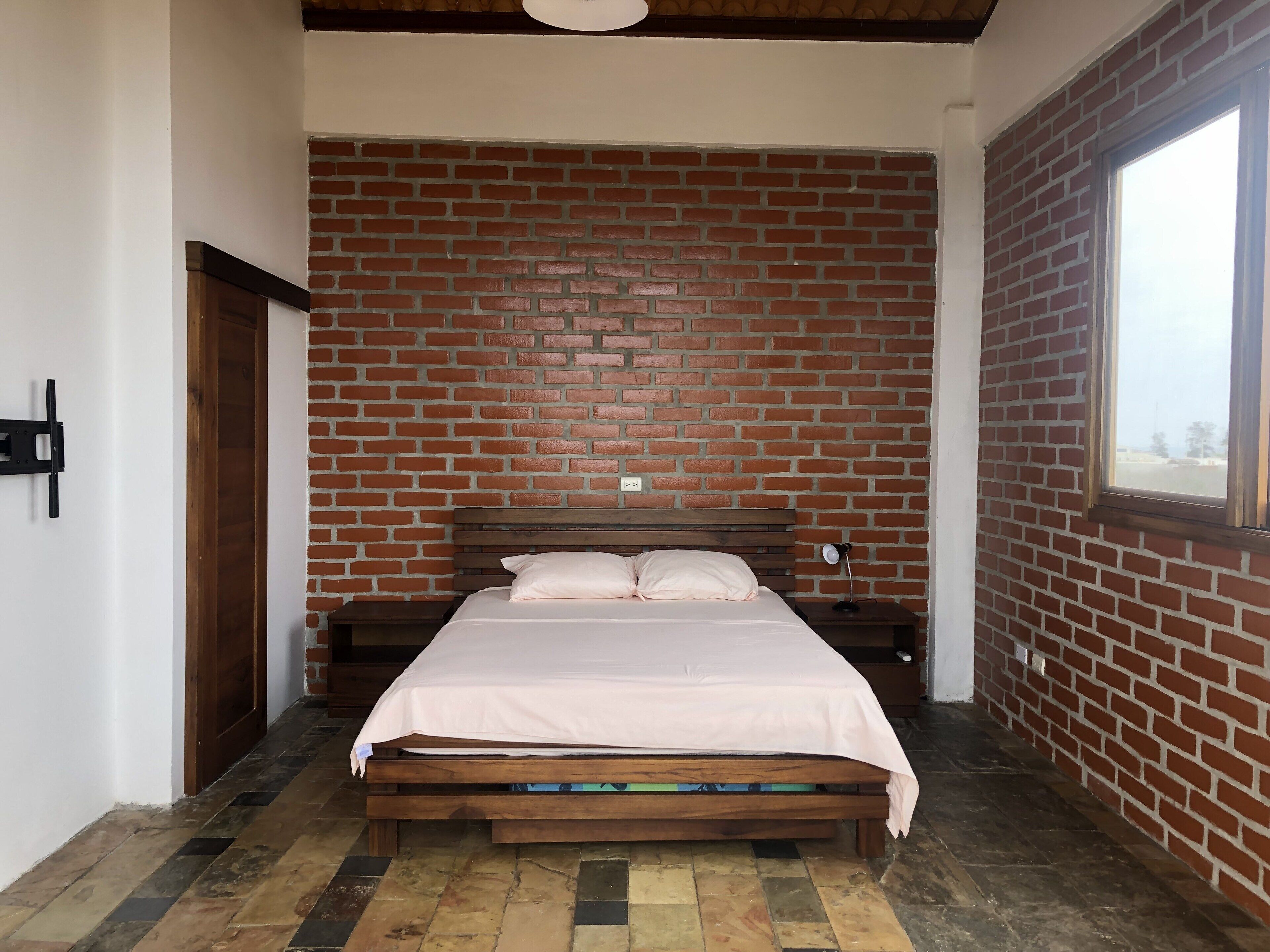 Un dormitorio con pared de ladrillo, estructura de cama de madera, mesita de noche y ventana.