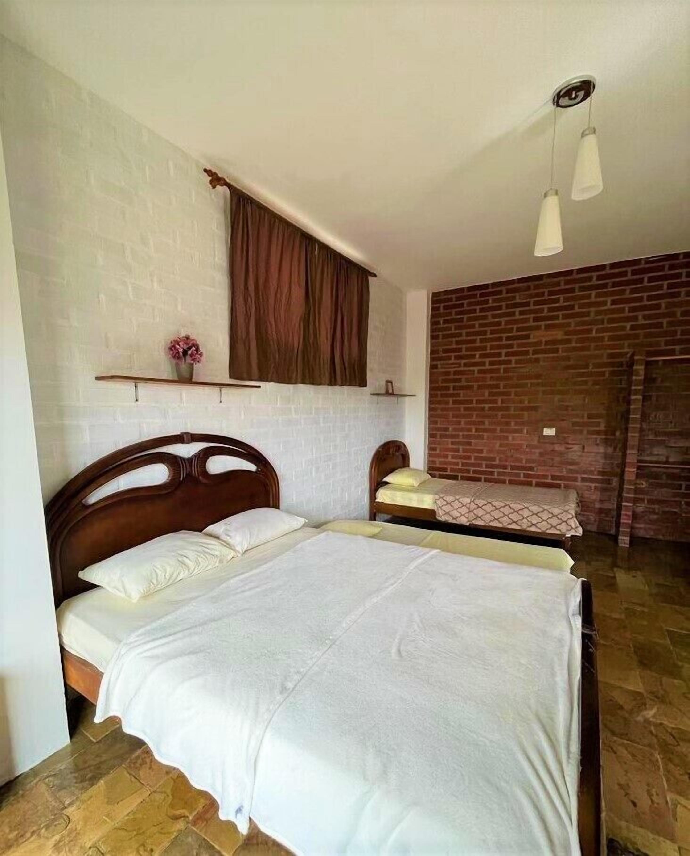 Una habitación con una cama grande, una cama más pequeña, una pared de ladrillo, un cabecero de madera, una ventana con cortina y una lámpara colgante.