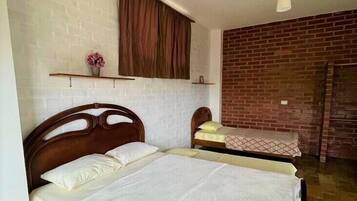 Una habitación con una cama grande, una cama más pequeña, una pared de ladrillo, un cabecero de madera, una ventana con cortina y una lámpara colgante.