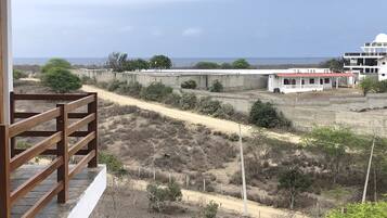 Vista desde un balcón sobre una zona costera con un sendero de tierra, vegetación escasa y algunos edificios a lo lejos.
