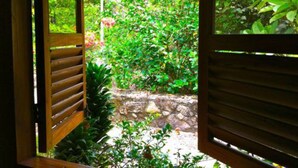 Une porte en bois, composée de lattes horizontales, donne sur un jardin luxuriant où poussent différentes espèces de plantes, et un mur de pierres en délimite le pourtour.