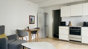 Ett modernt kök med vita skåp, ett kylskåp i rostfritt stål, en mikrovågsugn och en ugn. Det finns en matplats med en grå soffa och ett litet bord.