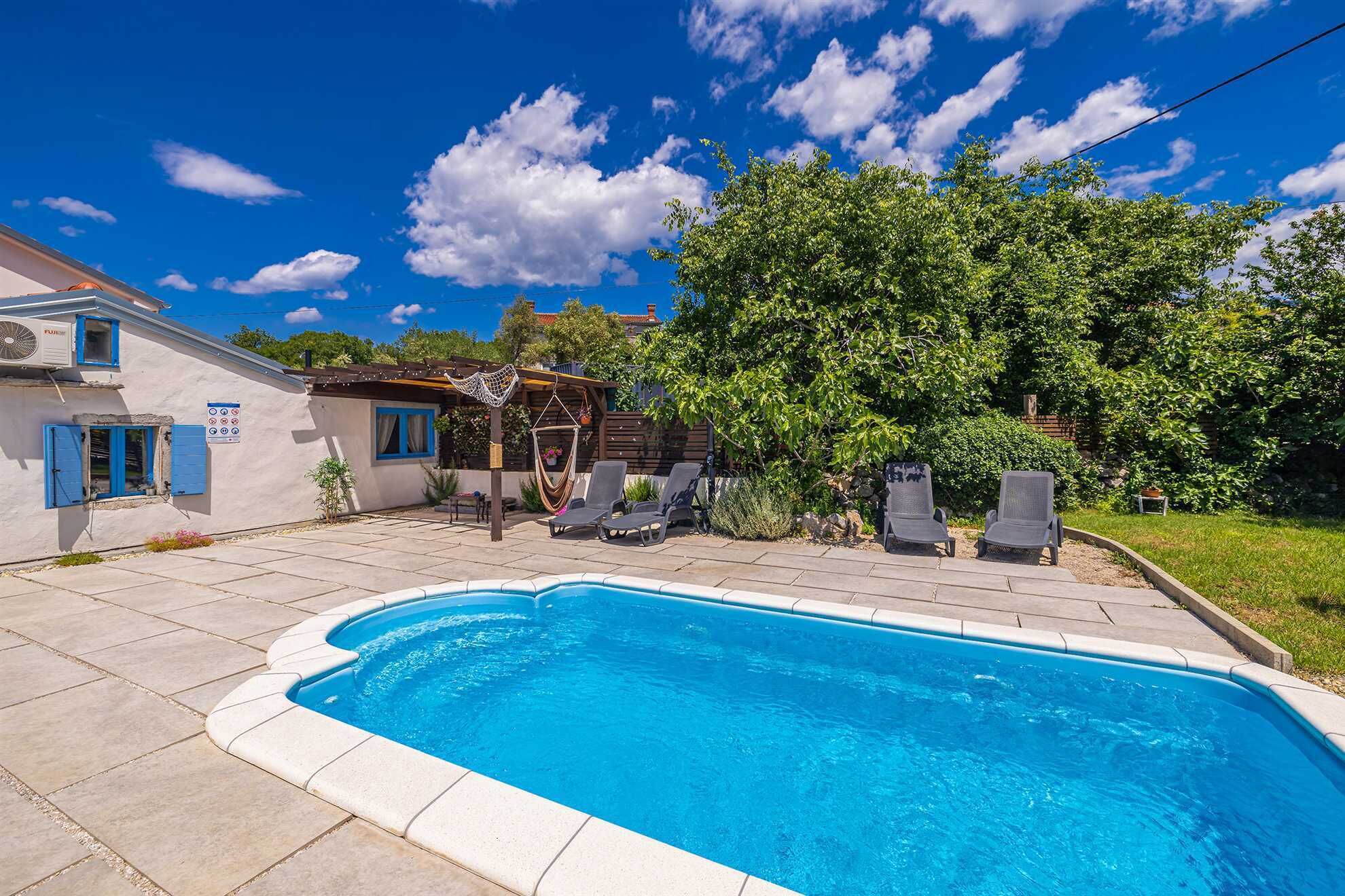 Une piscine avec des chaises longues, une pergola et une maison aux volets bleus.