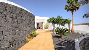 A pathway leading to a building with a stone wall, palm trees, and a clear sky. - Luxurious Villa with Panoramic View, Multiple Terraces and Large Pool (Costa Teguise)