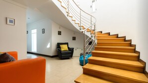A staircase with wooden steps and metal railings, a black leather chair with a yellow pillow, and an orange sofa in a well-lit room with white walls. - Luxurious Villa with Panoramic View, Multiple Terraces and Large Pool (Costa Teguise)