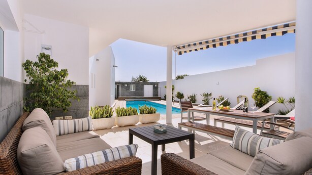 A sunlit outdoor patio with wicker furniture, a pool, and potted plants. - Luxurious Villa with Panoramic View, Multiple Terraces and Large Pool (Costa Teguise)