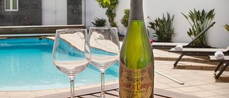 A bottle of wine and two glasses on a wooden table by a pool with a white wall and plants in the background.