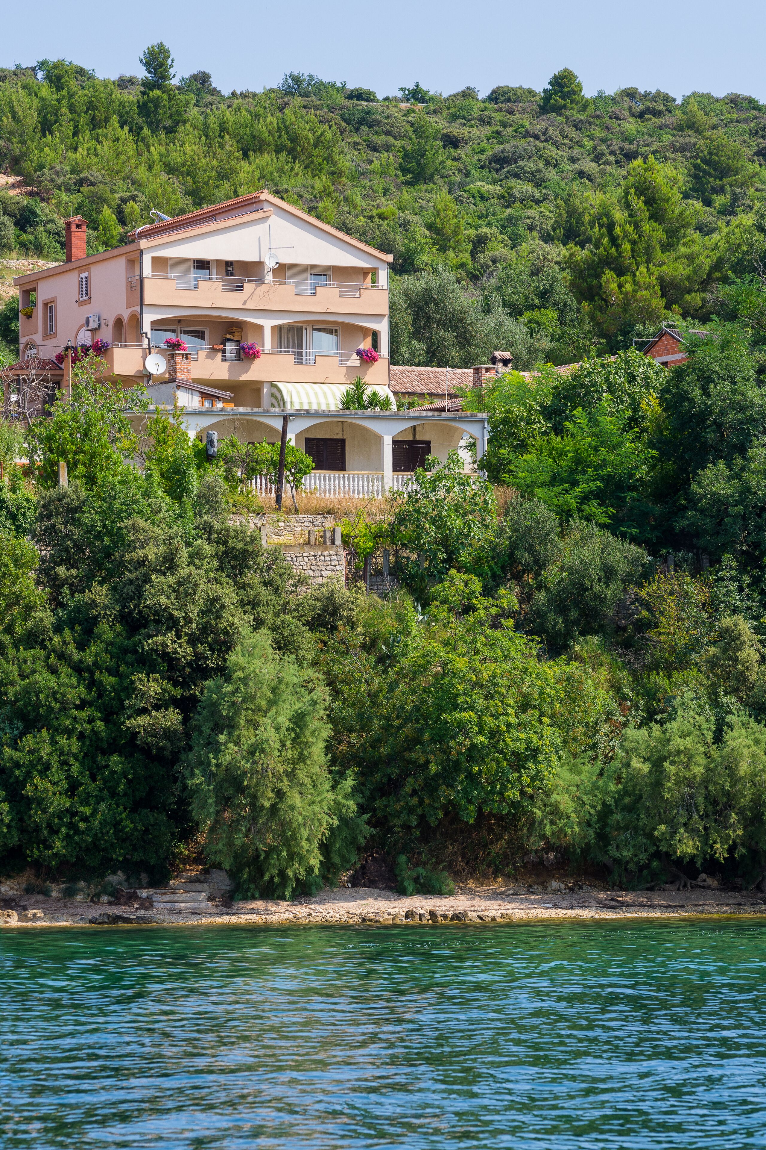 Une maison à deux étages dotée d’un balcon, entourée d’une végétation luxuriante et bordée d’un plan d’eau.