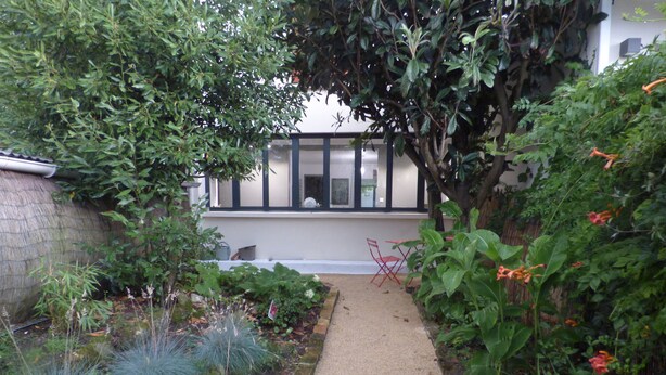 Un sendero en un jardín que lleva a un edificio con un ventanal con vistas al jardín.
