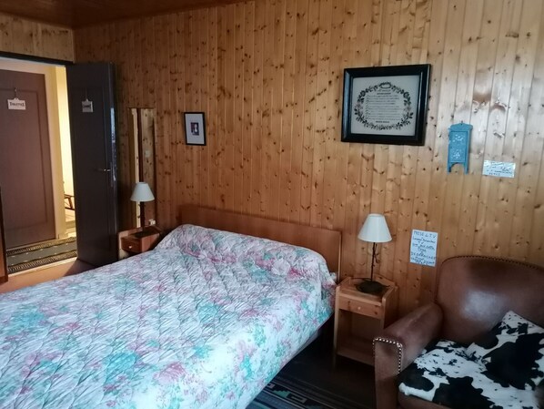 Une pièce aux murs lambrissés, contenant un lit, une chaise, une table de chevet et un certificat encadré accroché au mur.