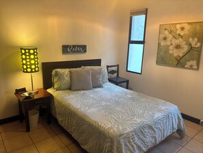 Ein Schlafzimmer mit einem Bett, einem Nachttisch, einer Lampe, einem Wandbild und einem Fenster.