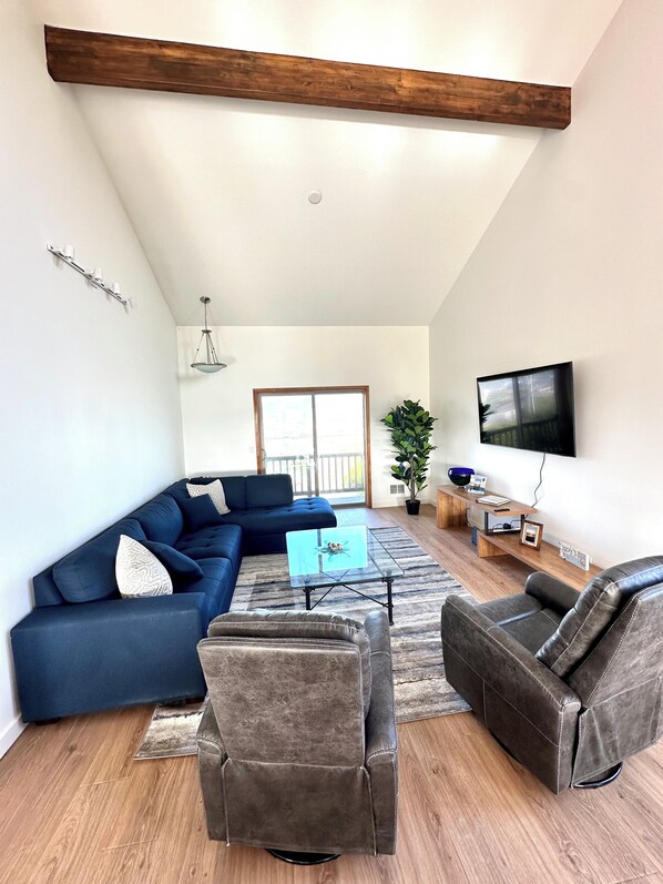 Ein Wohnzimmer mit einem blauen Modulsessel, zwei Ledersesseln, einem Glastisch, einer Holz-TV-Bank und einem großen Fenster mit Blick auf einen Balkon.