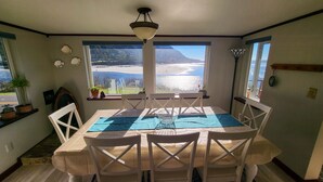 Een eethoek met een tafel en stoelen, grote ramen met uitzicht op een strand en bergen, en een plafondlamp.