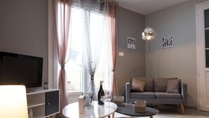 En moderne stue med et fladskærms-tv, et rundt sofabord, en grå sofa og et vindue med lette gardiner.