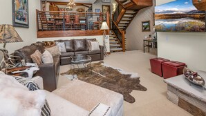 Un salon spacieux, doté d’un canapé en cuir, d’une table basse en verre et d’un grand tapis. Un escalier avec des rampes en bois mène à l’étage supérieur.