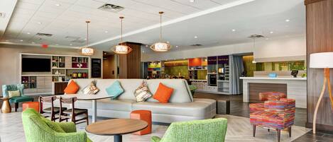 A modern hotel lobby with a sofa, chairs, a dining area, and a kitchen in the background.