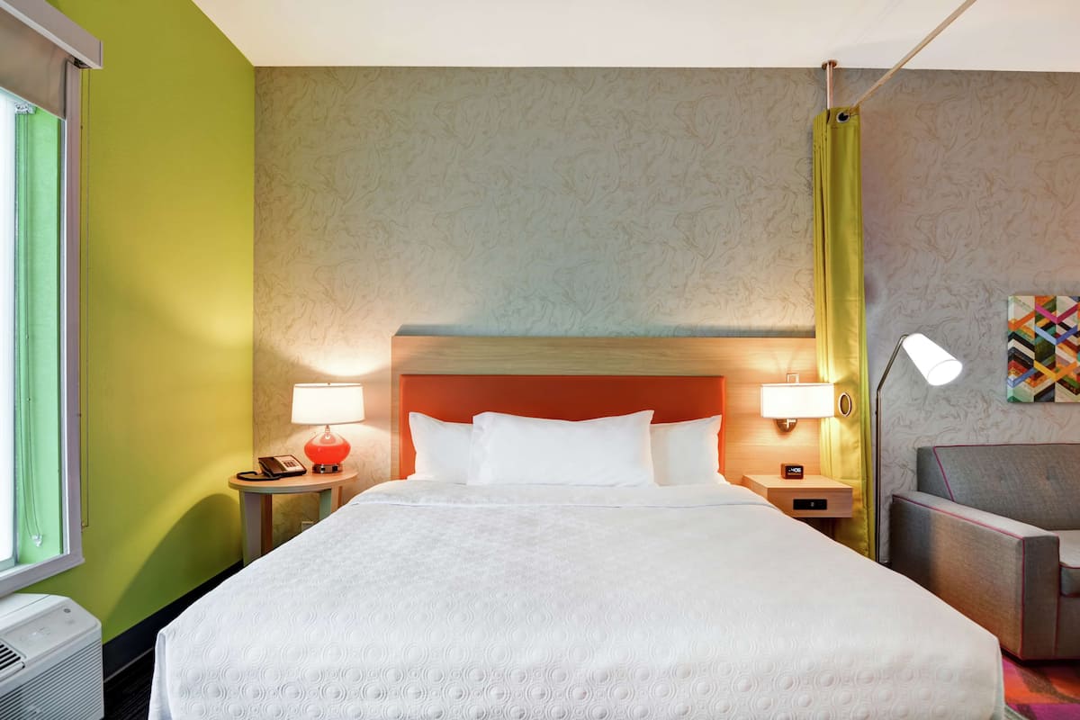 Habitación de hotel con una cama grande, dos mesitas de noche con lámparas, una silla y un televisor montado en la pared.