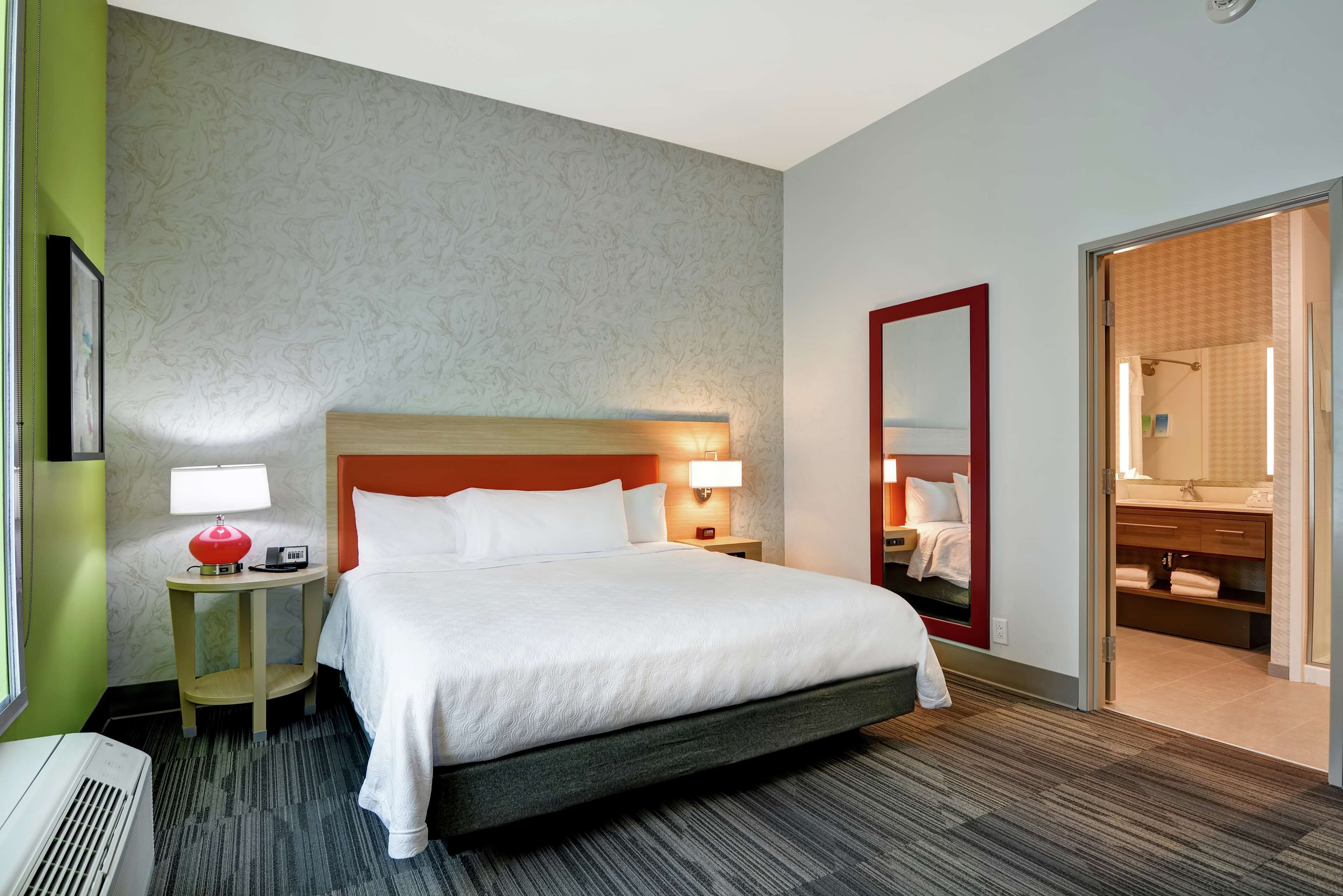 A hotel room with a large bed, a nightstand with a lamp, a mirror, and a bathroom visible through an open door.