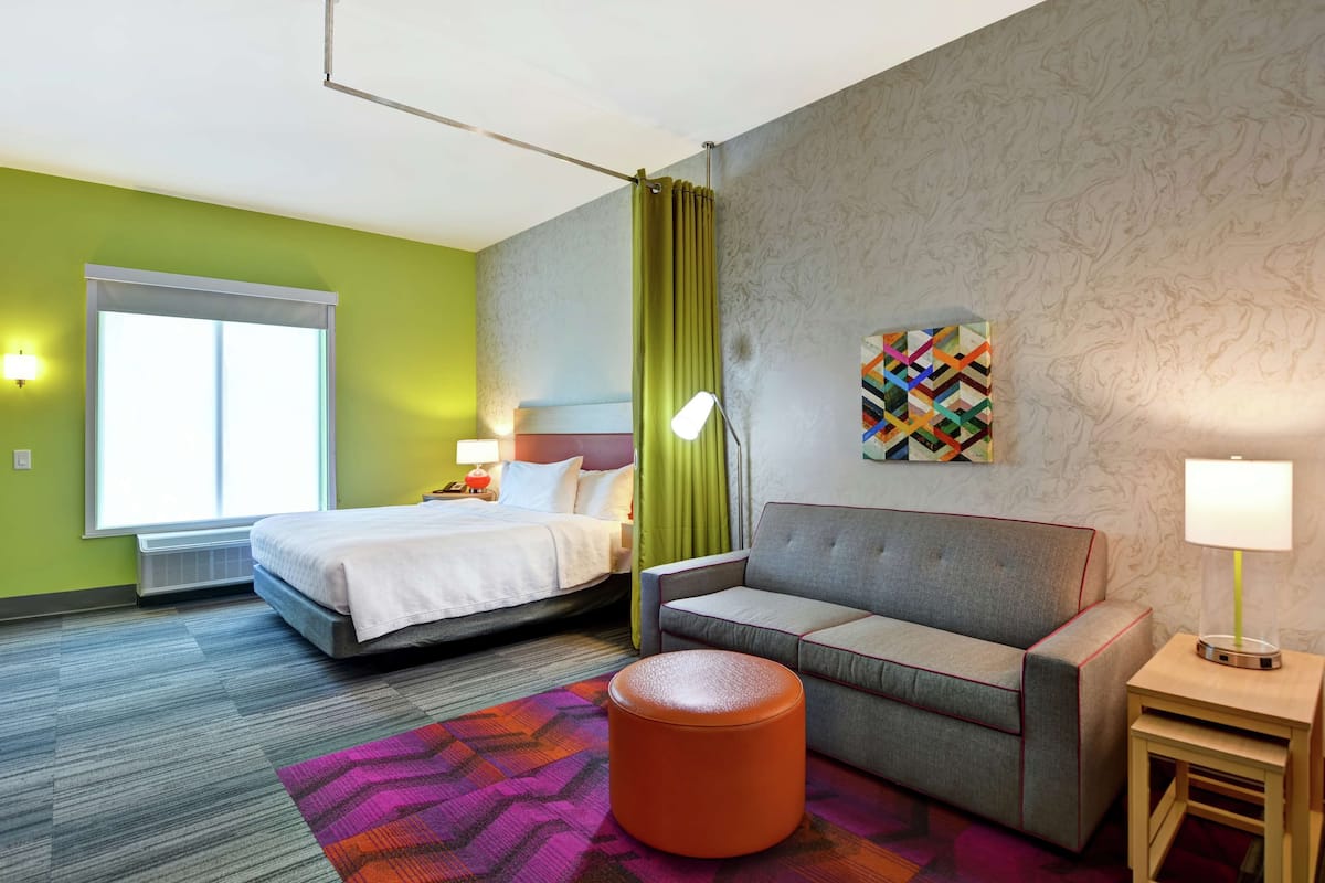 Habitación de hotel con cama, sofá, una mesita, lámpara, alfombra colorida y ventana con cortinas.
