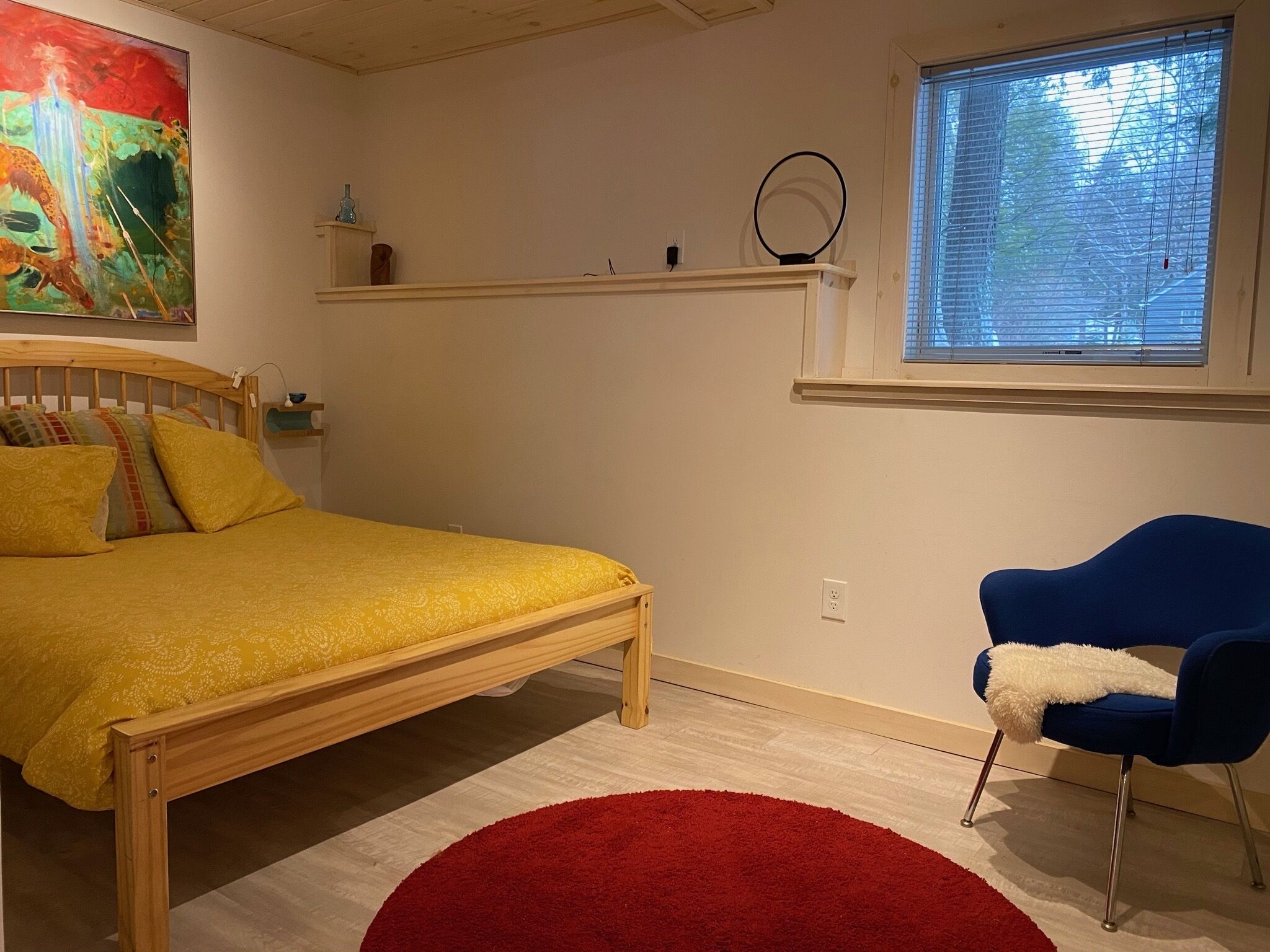 A bedroom with a wooden bed, a red rug, a blue chair, a painting, and a window with blinds.