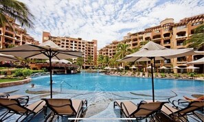 A hotel pool area with multiple lounge chairs and umbrellas, surrounded by multi-story buildings with balconies. - 5th Floor Wow views, BradAndJoeyRentals Closest to ocean - Villa La Estancia (Nuevo Vallarta)